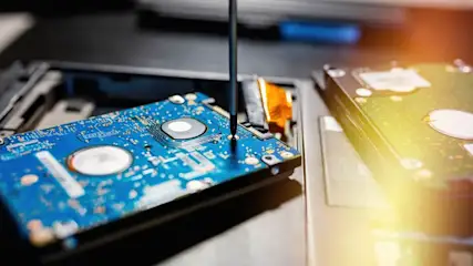 Nahaufnahme einer geöffneten Festplatte im Laptop, auf deren blauer Platine ein Schraubendreher ansetzt; daneben weitere Hardwarekomponenten unscharf.