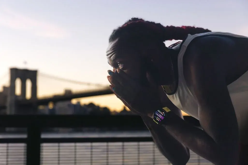 Person in weißem Tanktop mit Smartwatch und blauen Nägeln, vorgebeugt. Im Hintergrund verschwommen die Brooklyn Bridge bei Dämmerung.