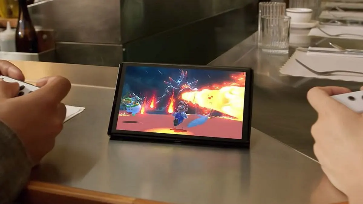 Zwei Personen spielen ein Videospiel auf einer Nintendo Switch auf einer Metalltheke. Das Spiel zeigt Luigi, ein Monster und Flammen. Küchenutensilien im Hintergrund.