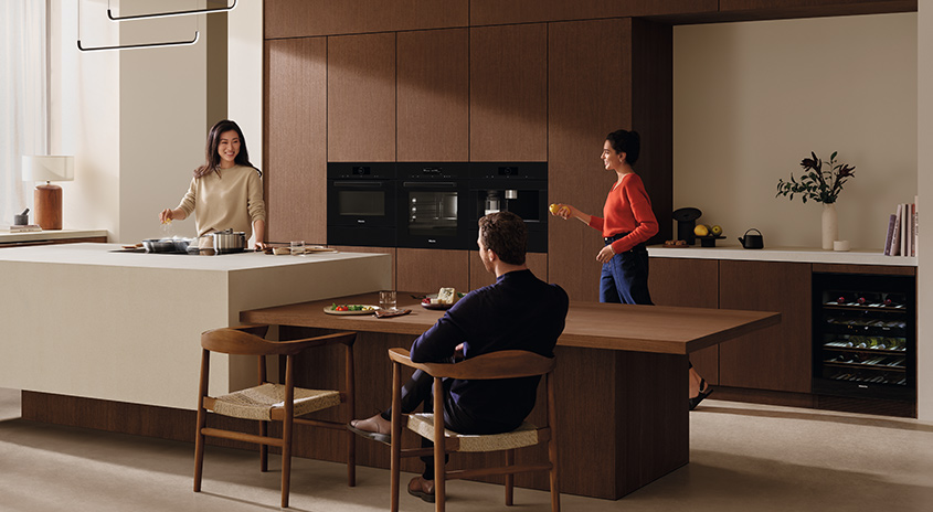 Eine Küche mit braunen Schränken und einer Kochinsel. Geräte sind eingebaut. Ein Holztisch steht im Raum.