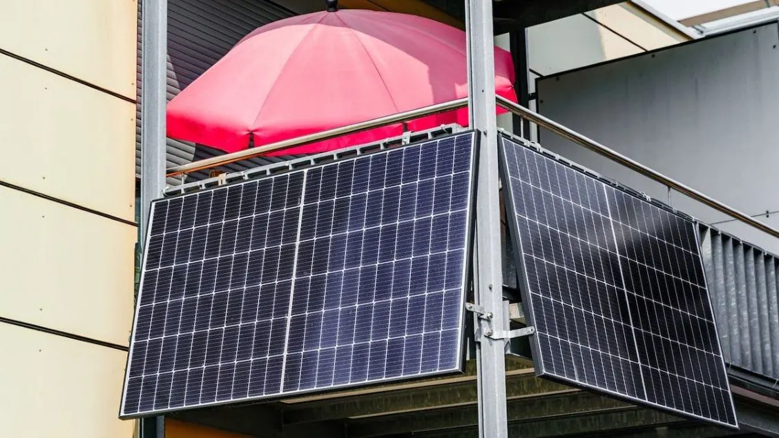Zwei dunkle Solarmodule sind an einem grauen Balkongeländer befestigt. Dahinter ist ein leuchtend roter Sonnenschirm und eine helle Gebäudewand.