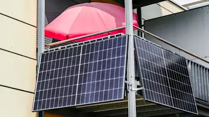 Zwei dunkle Solarmodule sind an einem grauen Balkongeländer befestigt. Dahinter ist ein leuchtend roter Sonnenschirm und eine helle Gebäudewand.