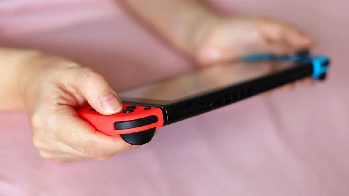 Hände halten eine schwarze Nintendo Switch Spielkonsole horizontal, mit rotem und blauem Controller, vor einem unscharfen rosa Hintergrund.