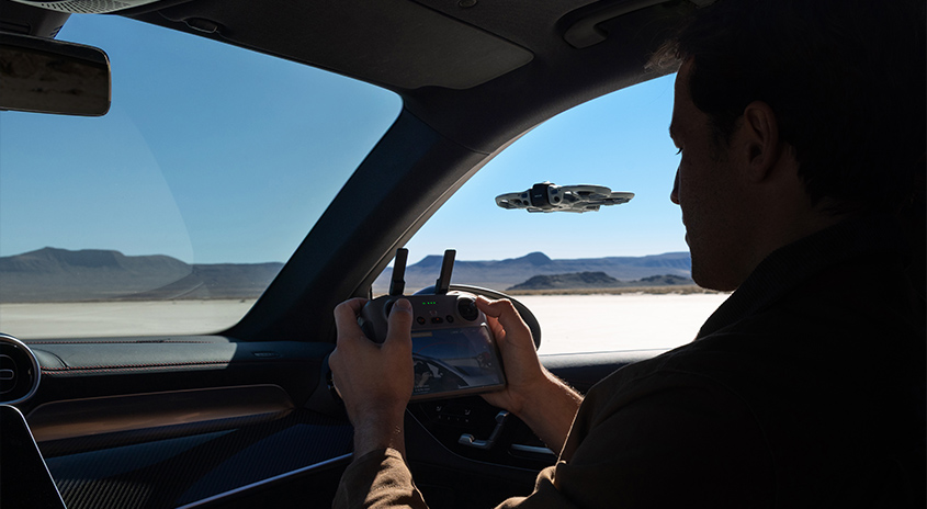 Person steuert DJI Drohne mit Controller aus dem Auto, durch das Seitenfenster ist die fliegende Drohne über heller Wüstenlandschaft zu sehen.