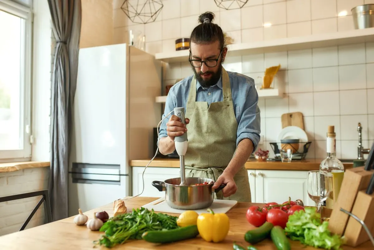 Ein Mann mit Bart und Brille benutzt einen Stabmixer in einem Topf, er befindet sich dabei in einer Küche, Gemüse liegt rund um den topf auf der Arbeitsplatte.