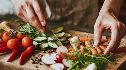 Hände schneiden Zucchini auf einem Holzbrett mit frischem Gemüse wie Tomaten, Möhren und Radieschen.