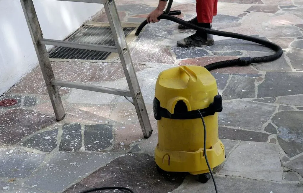Person saugt weißen Schutt mit gelbem Kärcher Staubsauger von mehrfarbigem Steinboden; daneben steht eine silberne Leiter.