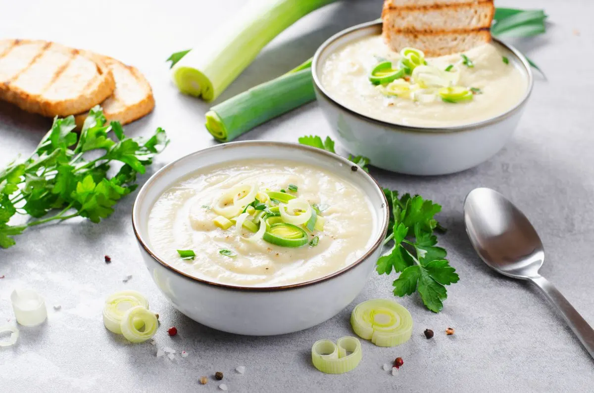 Kartoffel Lauch Creme Suppe in 2 Tellern, mit Lauch ringen als Topping, daneben liegen Petersil, Lauch und getostetes Brot