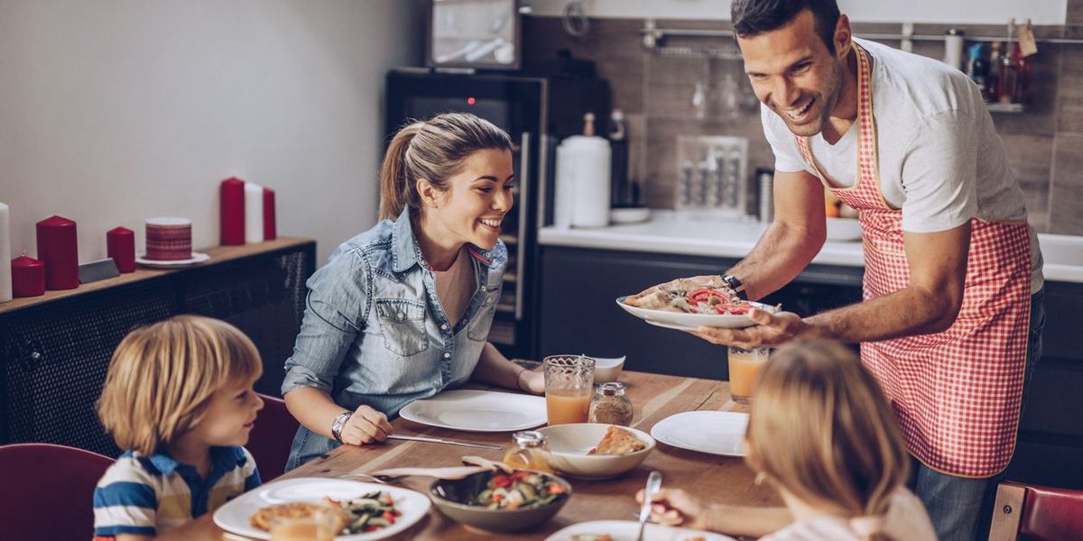Ein Mann in rotkarierter Kochschürze serviert Essen für eine lächelnde Familie.