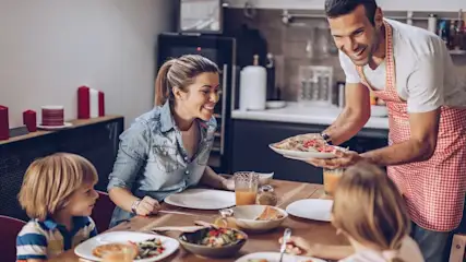 Ein Mann in rotkarierter Kochschürze serviert Essen für eine lächelnde Familie.