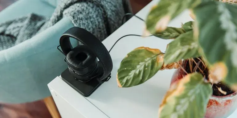 Schwarze Kopfhörer auf einer Ladestation auf einem weißen Tisch neben einer Pflanze mit grünen und braunen Blättern in einem roten Topf.