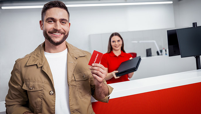 Ein Mann hält eine rote Karte vor einem roten Tresen. Im Hintergrund eine Frau mit Laptop und Monitore.
