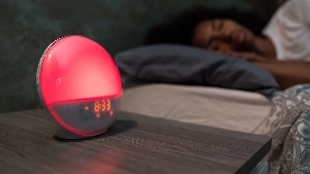 Roter Lichtwecker auf dunklem Nachttisch zeigt 8:33. Im Hintergrund schläft eine Person auf einem grauen Kissen im Bett.