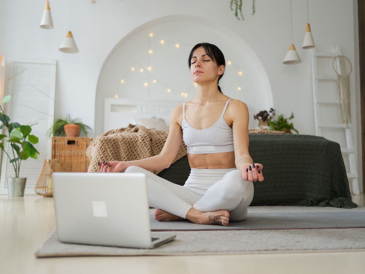 Eine Person in heller Sportkleidung meditiert mit geschlossenen Augen auf einer Yogamatte vor einem Laptop. Im Hintergrund ein Bett, Pflanzen und Lichter.