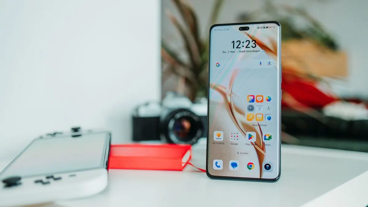 Smartphone zeigt 12:23 Uhr und Apps. Daneben ein weißer Handheld mit roter Powerbank und eine Kamera im Hintergrund.