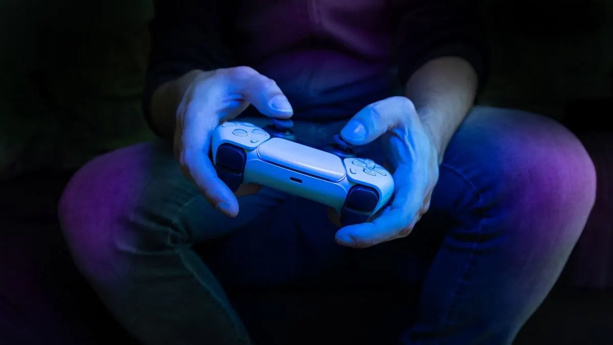 Nahaufnahme von Händen, die einen weißen PlayStation Controller halten, bläuliche Beleuchtung.