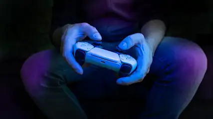 Nahaufnahme von Händen, die einen weißen PlayStation Controller halten, bläuliche Beleuchtung.