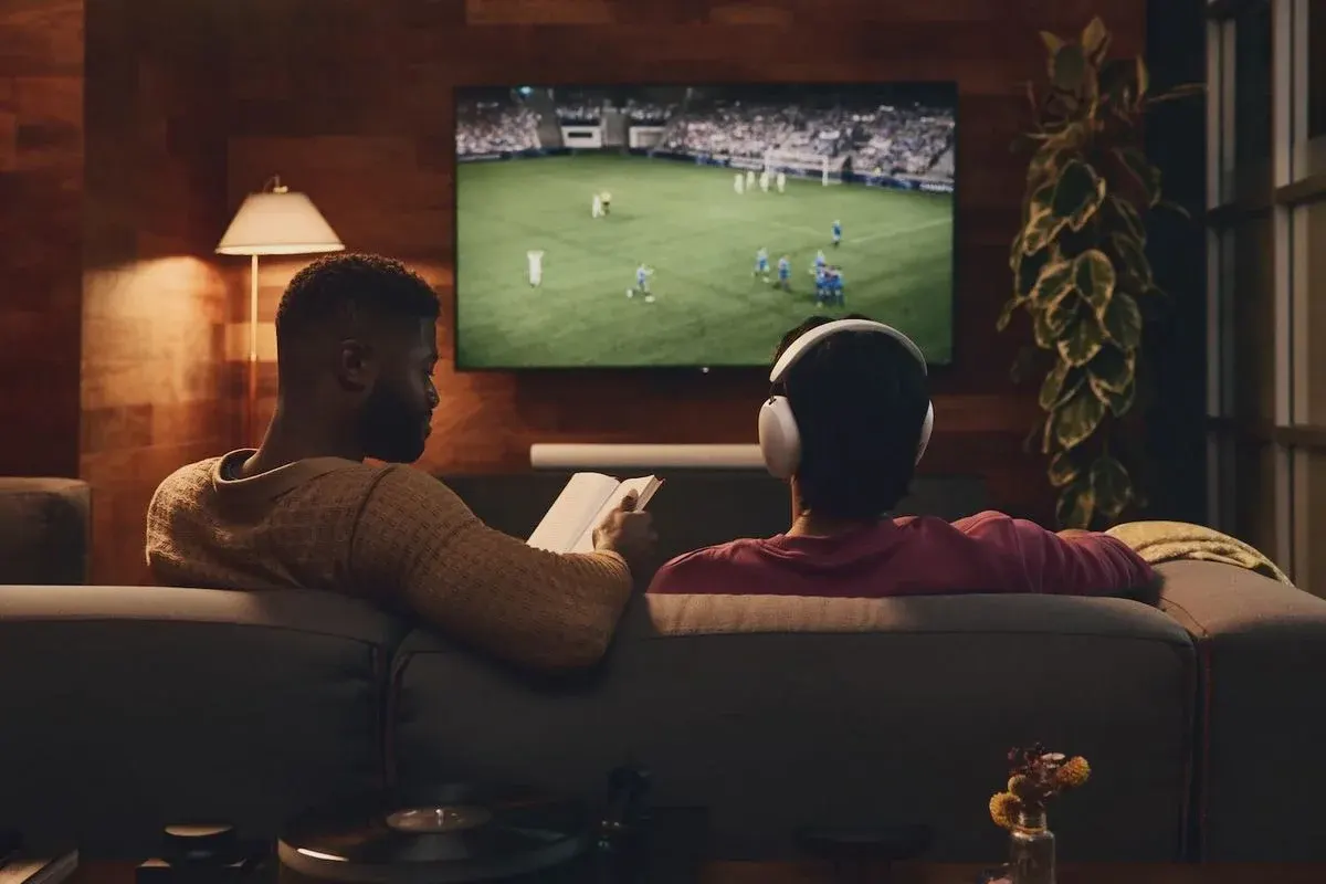 Zwei Menschen auf einem Sofa schauen gemeinsam ein Fußballspiel im Fernsehen, während eine Person ein Tablet hält und die andere Kopfhörer trägt.
