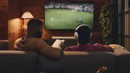 Zwei Menschen auf einem Sofa schauen gemeinsam ein Fußballspiel im Fernsehen, während eine Person ein Tablet hält und die andere Kopfhörer trägt.
