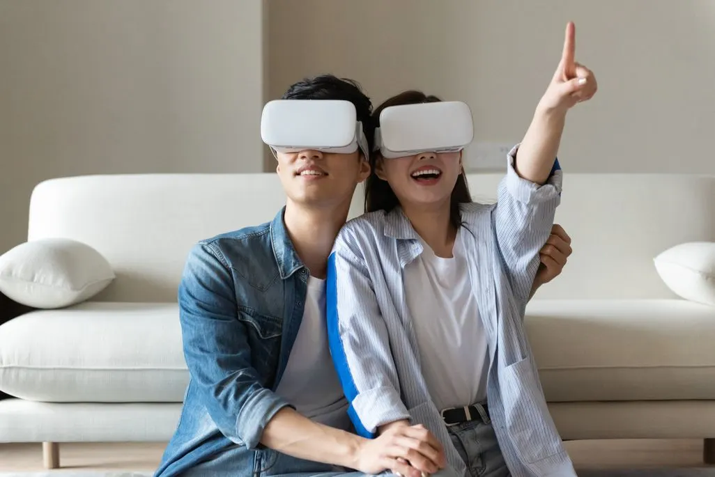Zwei Personen sitzen auf dem Boden mit VR-Brillen und zeigen in die Luft.