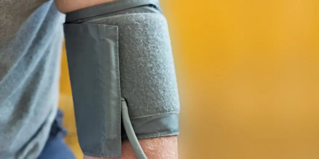 Ein Arm trägt eine graue Blutdruckmanschette über einem grauen Hemd vor einem unscharfen, orange-gelben Hintergrund.