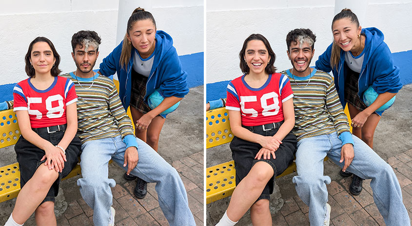 Drei Personen sitzen auf einer gelben Bank vor einer weißen und blauen Wand. Zwei sitzen, eine steht, alle lächeln.