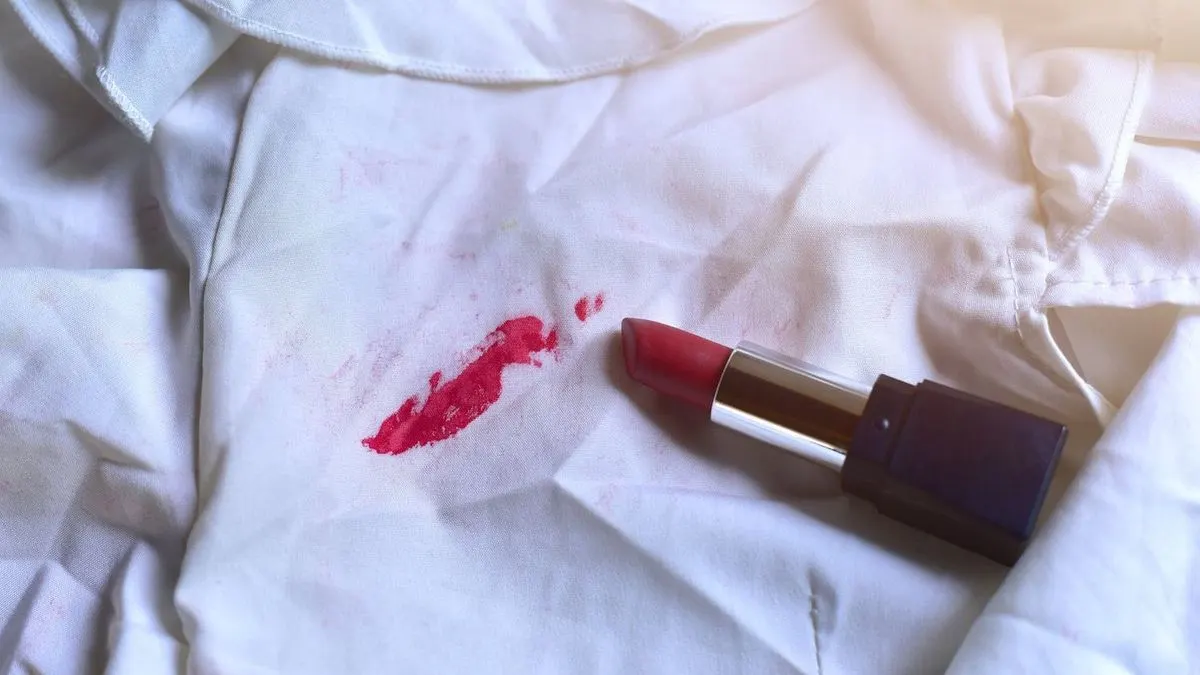 Offener roter Lippenstift liegt neben einem Lippenstift-Fleck auf weißem Stoff.