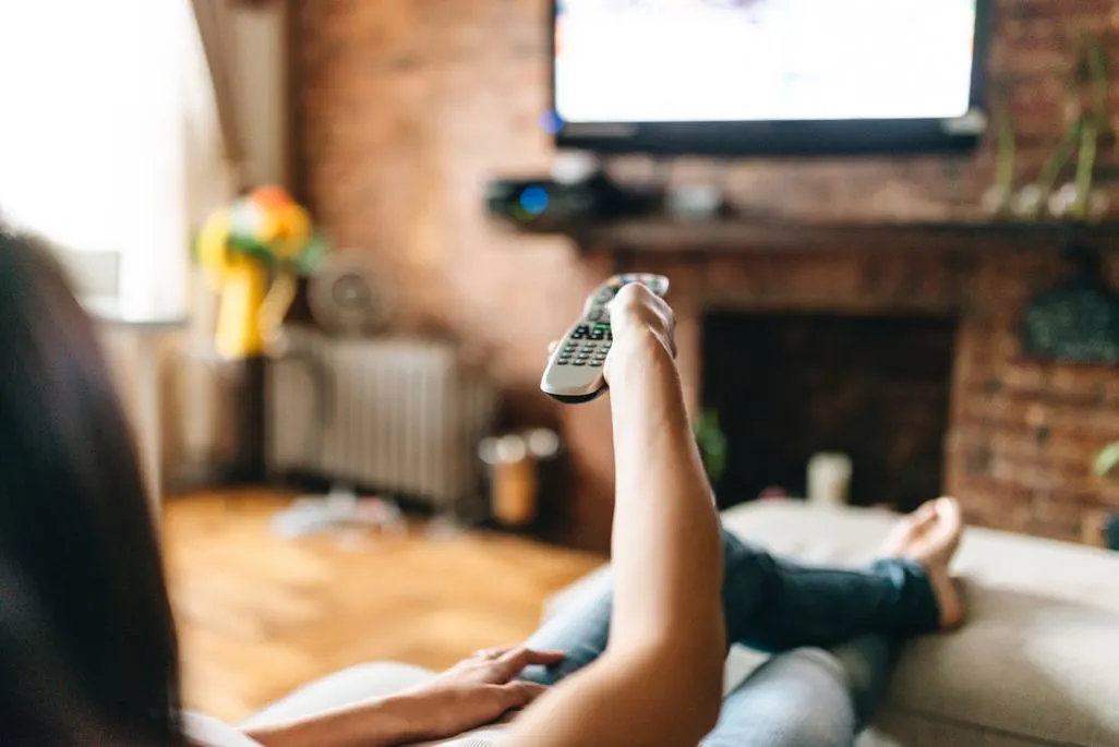 Person liegt auf dem Sofa und richtet eine Fernbedienung auf den Fernseher.