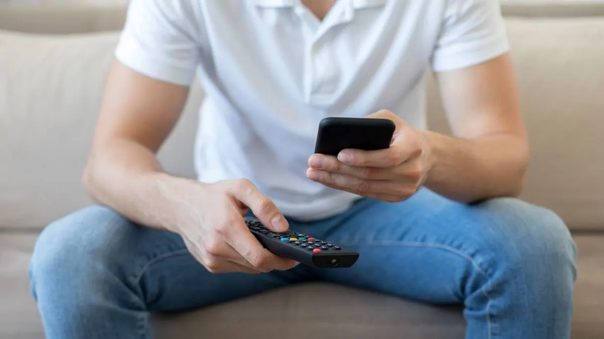 Mann sitzt auf dem Sofa und hält eine Fernbedienung und ein Smartphone in den Händen.