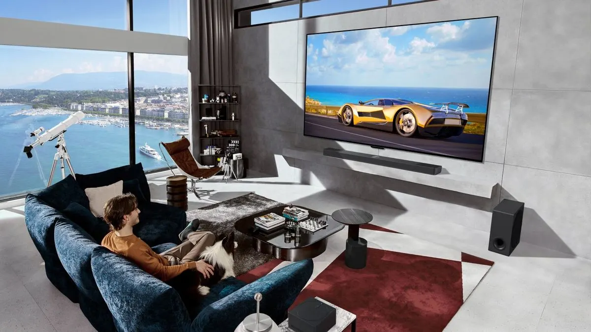 Eine Person sitzt mit einem Hund auf einem Sofa und schaut einen großen Fernseher.