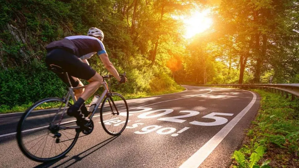 Radfahrer mit Helm fährt auf einer kurvigen Landstraße durch einen sonnigen Wald. Auf der Straße steht groß "2025 let's go".