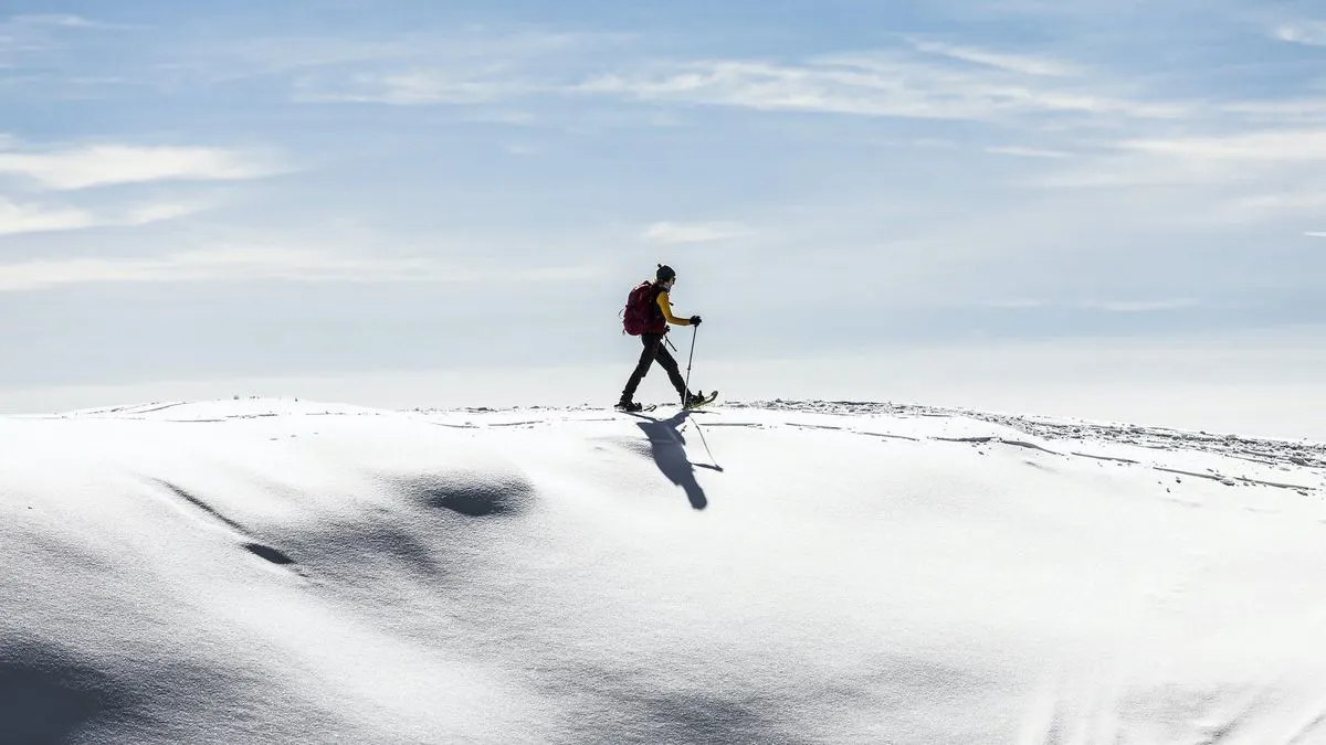 Person in Gelb mit rotem Rucksack geht auf Schneeschuhen über Schnee. Blauer Himmel mit Wolken und fernen Schneehügeln.