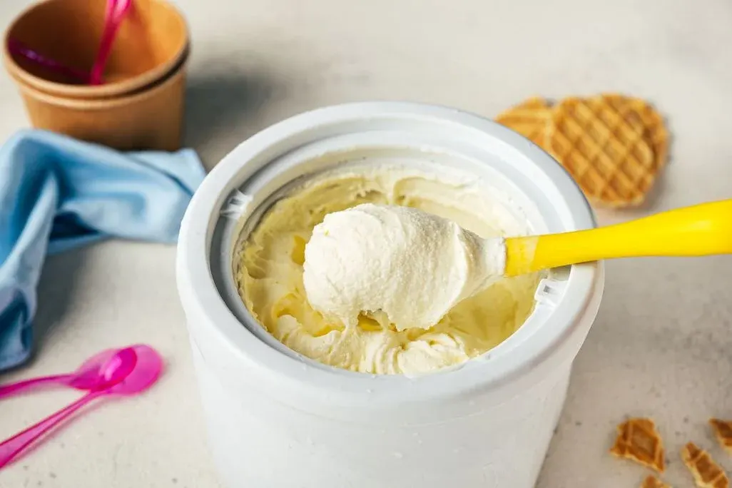 Vanilleeis wird mit gelbem Löffel aus weißer Eismaschine portioniert. Im Hintergrund farbige Eislöffel, bunte Sorbets und Waffelkegel auf hellem Untergrund.