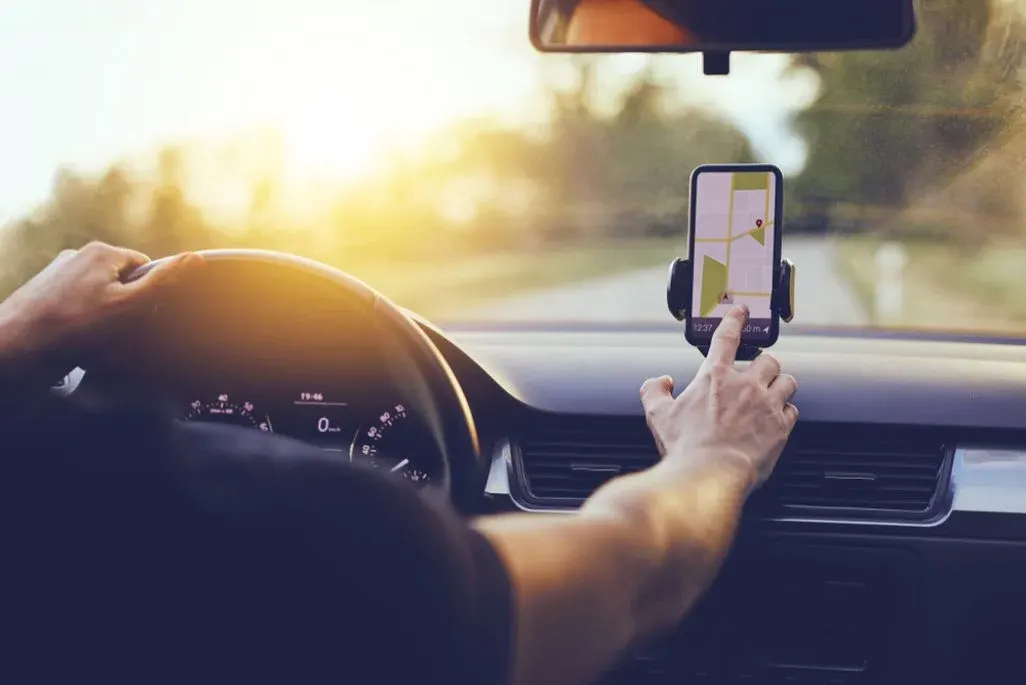 Person fährt Auto, Hände am Lenkrad und berühren Smartphone mit Navigationskarte. Helle Sonne über grüner Landschaft.