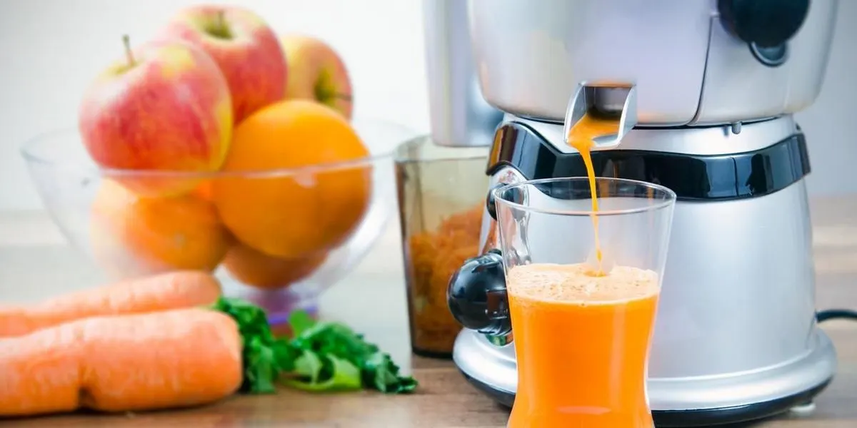 Ein Entsafter füllt ein Glas mit frisch gepresstem Karottensaft, daneben liegen Äpfel, Orangen und Karotten