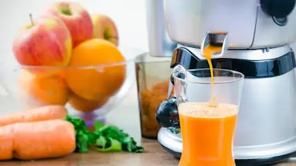 Ein Entsafter füllt ein Glas mit frisch gepresstem Karottensaft, daneben liegen Äpfel, Orangen und Karotten