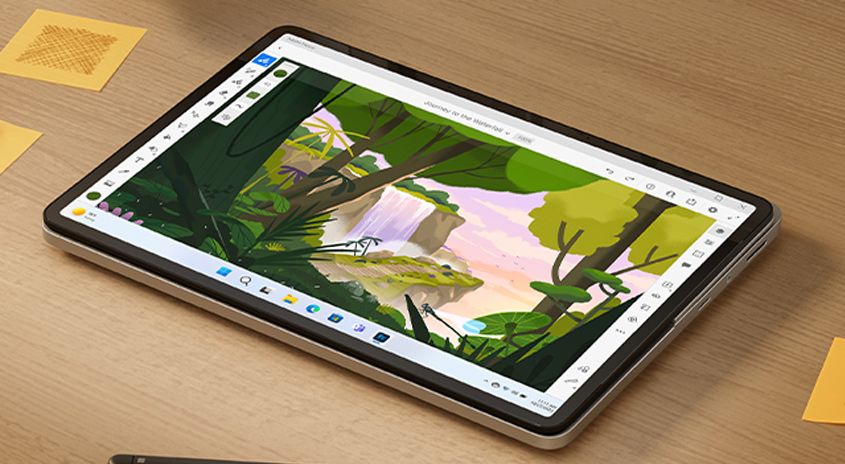 Ein Tablet zeigt eine digitale Zeichnung einer Dschungelszene mit Wasserfall, Bäumen und Teichen auf einem Holztisch mit Notizen.