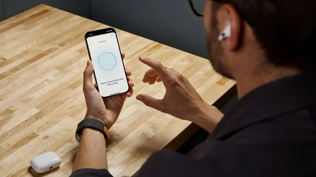 Mann mit Airpods im Ohr sitzt an einem Holztisch und nutzt sein Smartphone.