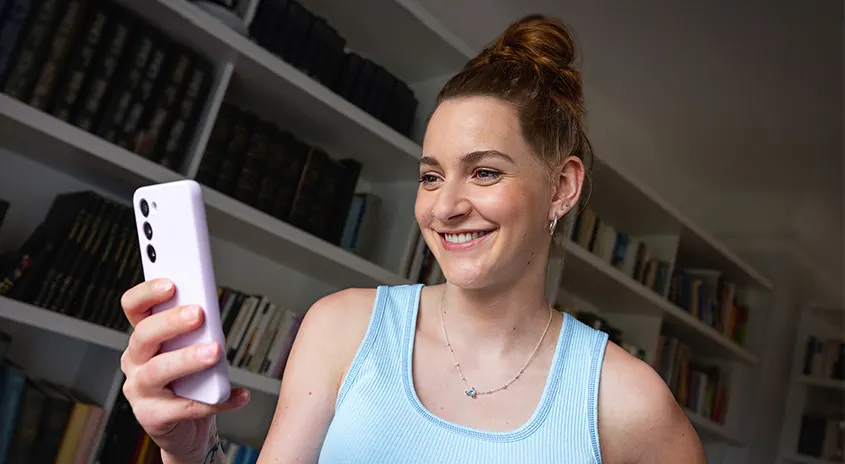 Eine Person hält ein lila Smartphone vor einem Bücherregal. Das Regal ist mit zahlreichen Büchern gefüllt.