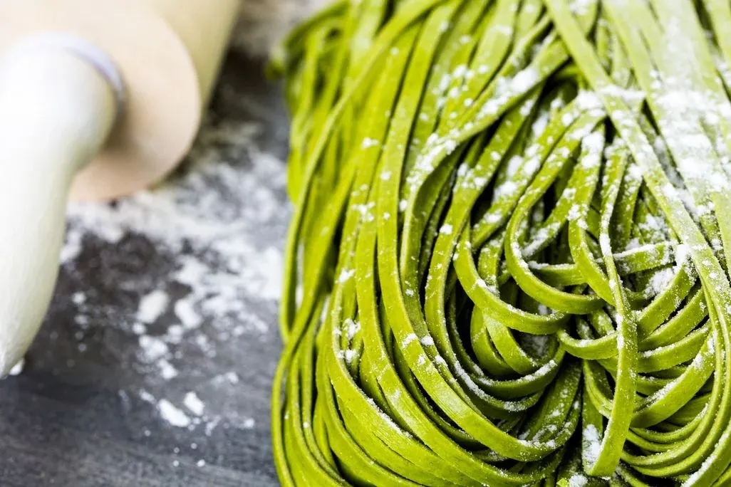 Haufen grüne, ungekochte Pasta, mit Mehl bestäubt, liegt auf dunkler Fläche neben einem Nudelholz mit Mehlspuren.