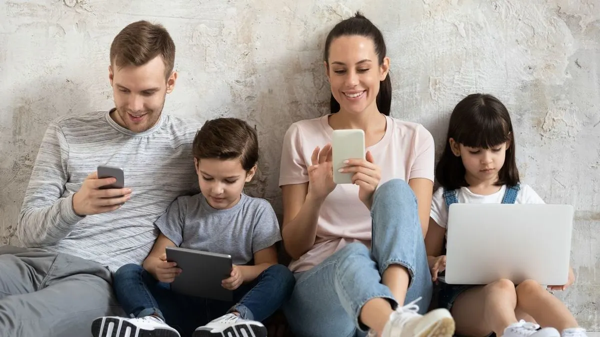Familie sitzt vor heller Wand: Mann mit grauem Smartphone, Junge mit Tablet, Frau mit hellem Smartphone, Mädchen mit weißem Laptop.