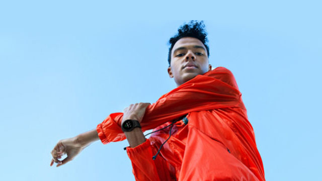 Ein Mann in oranger Jacke dehnt sich vor blauem Himmel. Er trägt eine Armbanduhr.