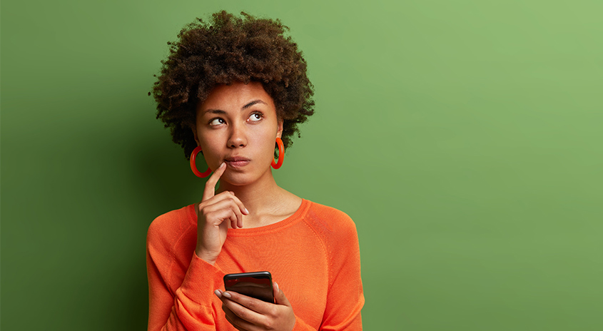 Eine Frau hält ein Smartphone, denkt nach, mit Finger am Kinn, vor grünem Hintergrund.