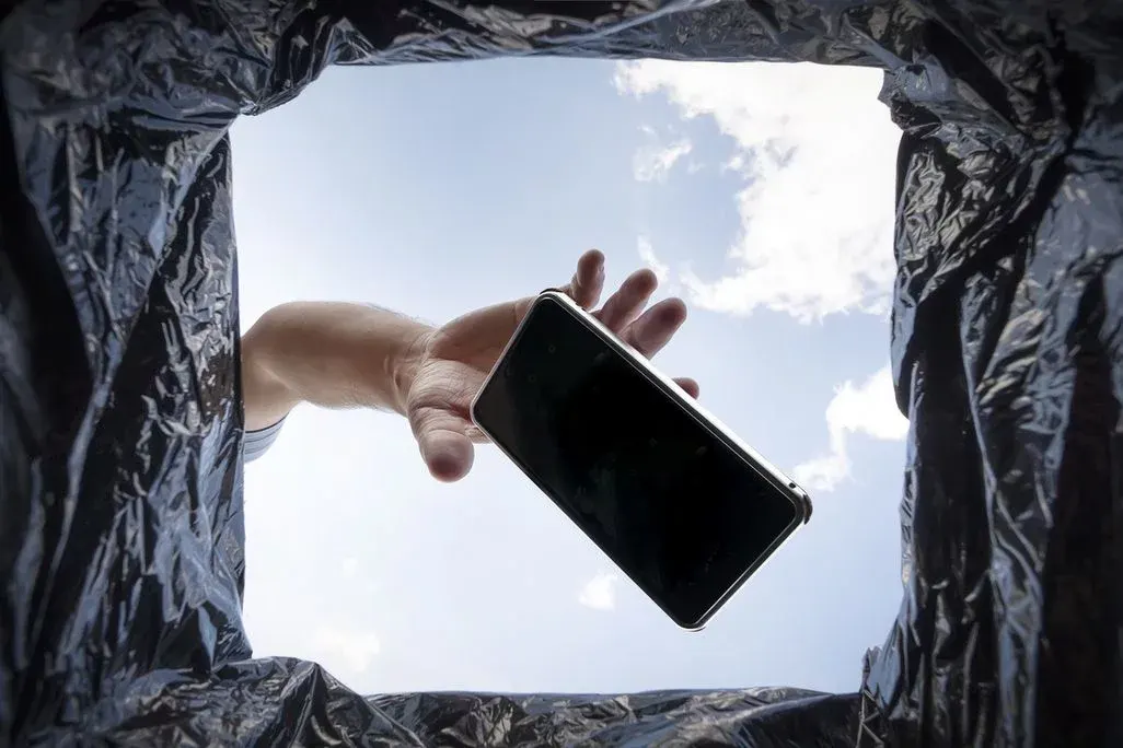 Blick aus einem Mülleimer: Eine Hand lässt ein Smartphone in einen schwarzen Müllsack fallen – Himmel im Hintergrund.