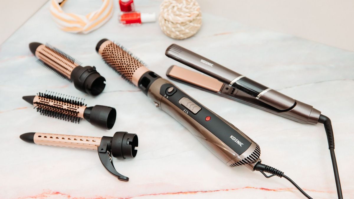 Bronze-schwarze Haarstyling-Tools mit Aufsätzen und ein Glätteisen auf Marmor. Ein Stirnband und Nagellack sind im Hintergrund.