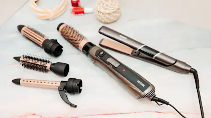 Bronze-schwarze Haarstyling-Tools mit Aufsätzen und ein Glätteisen auf Marmor. Ein Stirnband und Nagellack sind im Hintergrund.