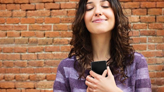 Eine Frau hält ein schwarzes Smartphone vor einer roten Backsteinmauer und lächelt. Sie trägt einen lila Pullover.