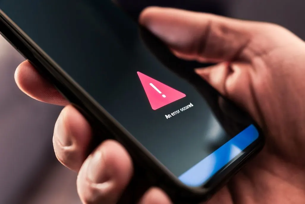 Eine Hand hält ein schwarzes Smartphone. Der Bildschirm zeigt ein rotes Warndreieck mit Ausrufezeichen und dem Text „An error occurred“. Eine blaue Reflexion ist unten sichtbar.