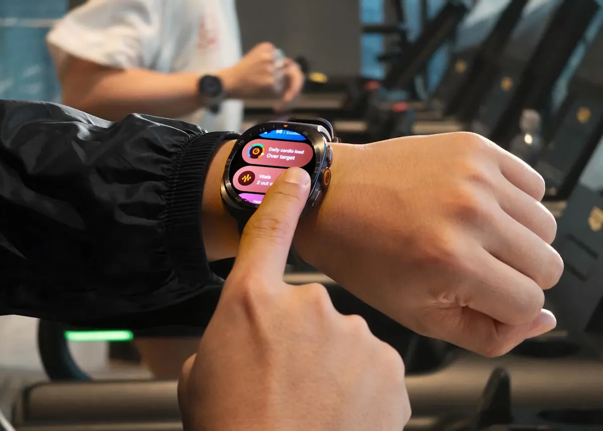 Eine Person tippt mit dem Finger auf die Samsung Galaxy Ultra2 auf ihrem Handgelenk. Im Hintergrund ist ein Fitnesscenter zu sehen.