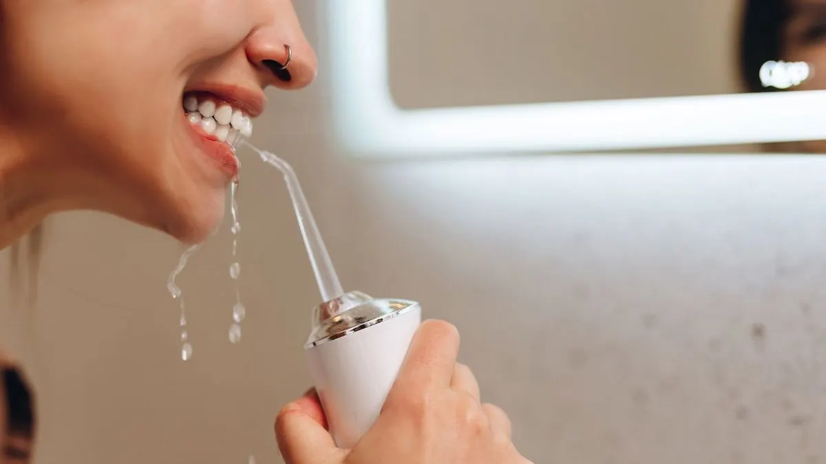 Person nutzt weiße Munddusche; Wasser spritzt aus dem Mund. Hand hält Gerät. Im Hintergrund ist ein beleuchteter Spiegel.